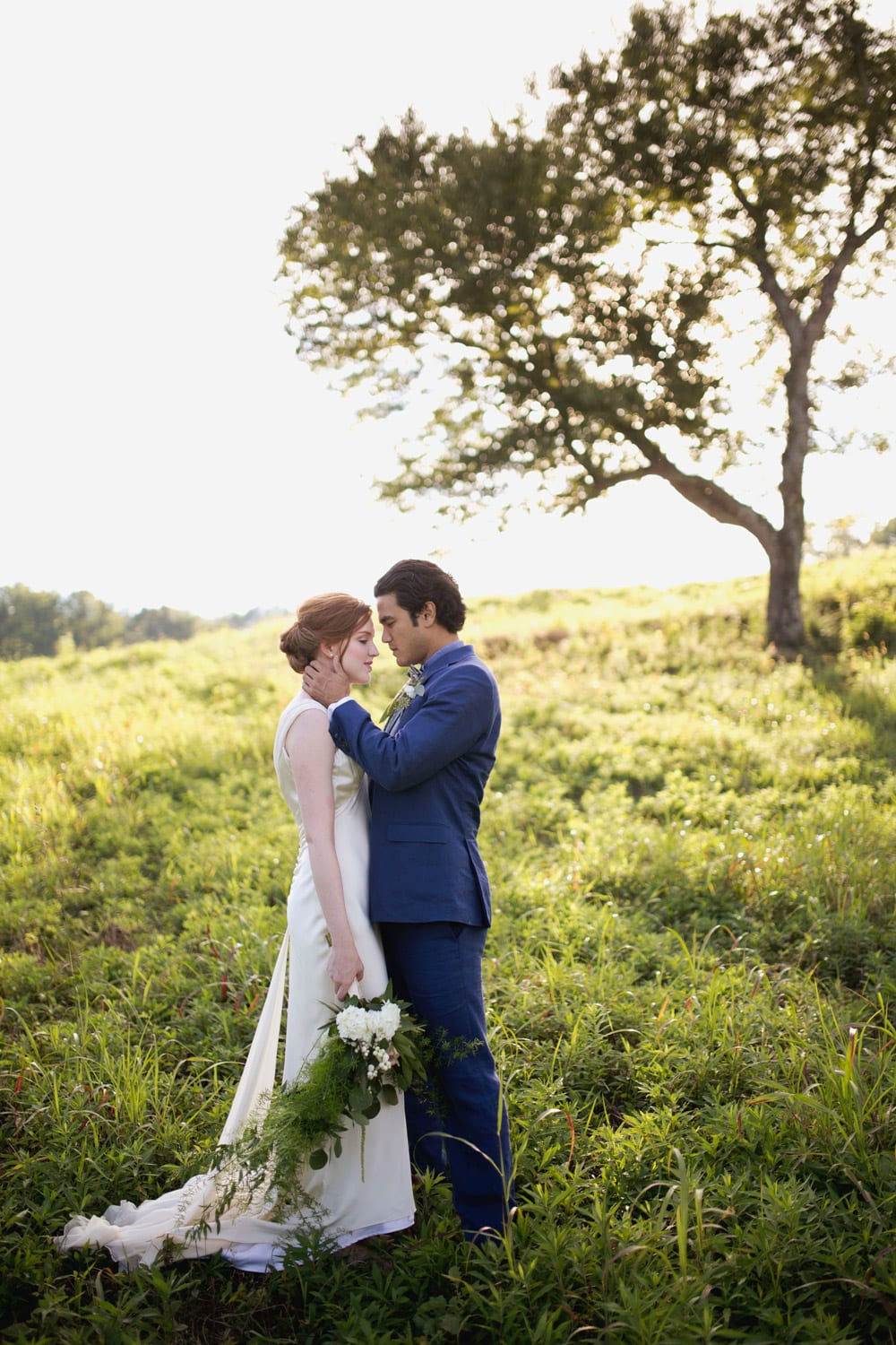 Groom holds bride in front of tree in the pasture