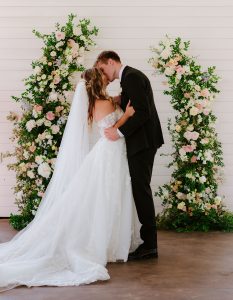 Bride and Groom kiss each other in front of floral decorations and a white wooden wall at a north Georgia wedding venue