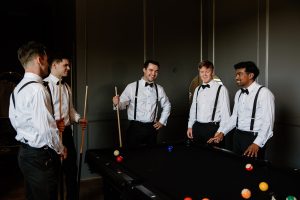 Groom and groomsmen stand around billiards table playing pool and smiling at The Greystone Estate wedding venue