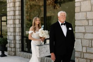Bride walks behind father with flowers and presents in front of The Grand wedding venue in north Georgia