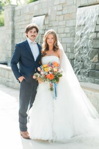 Bride and groom pose with flowers in front of grey wall with waterfall at a north Georgia wedding venue