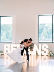 Groom leans bride back and kisses her while standing in front of a sign with their last name at a wedding venue in North Georgia